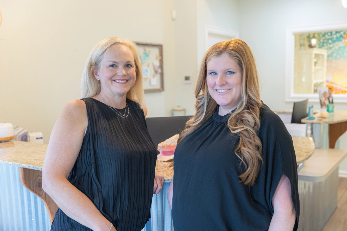 Two women in black attire smile at camera in a modern dental office interior.