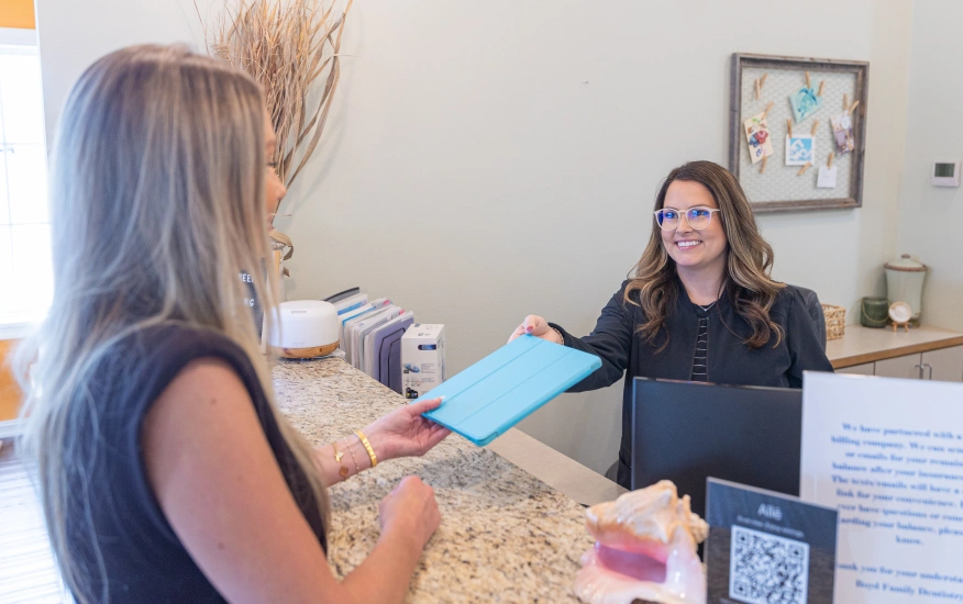 Woman holding blue folder speaking with receptionist at dental office front desk.