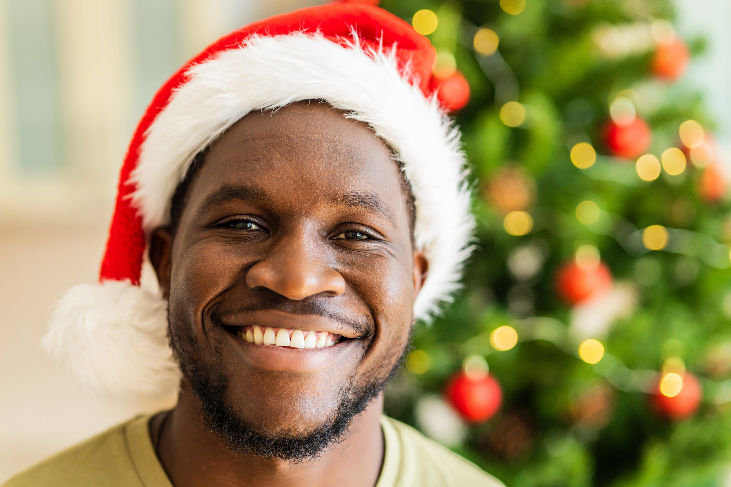 Man wearing Santa hat smiling at camera with decorated Christmas tree in background.